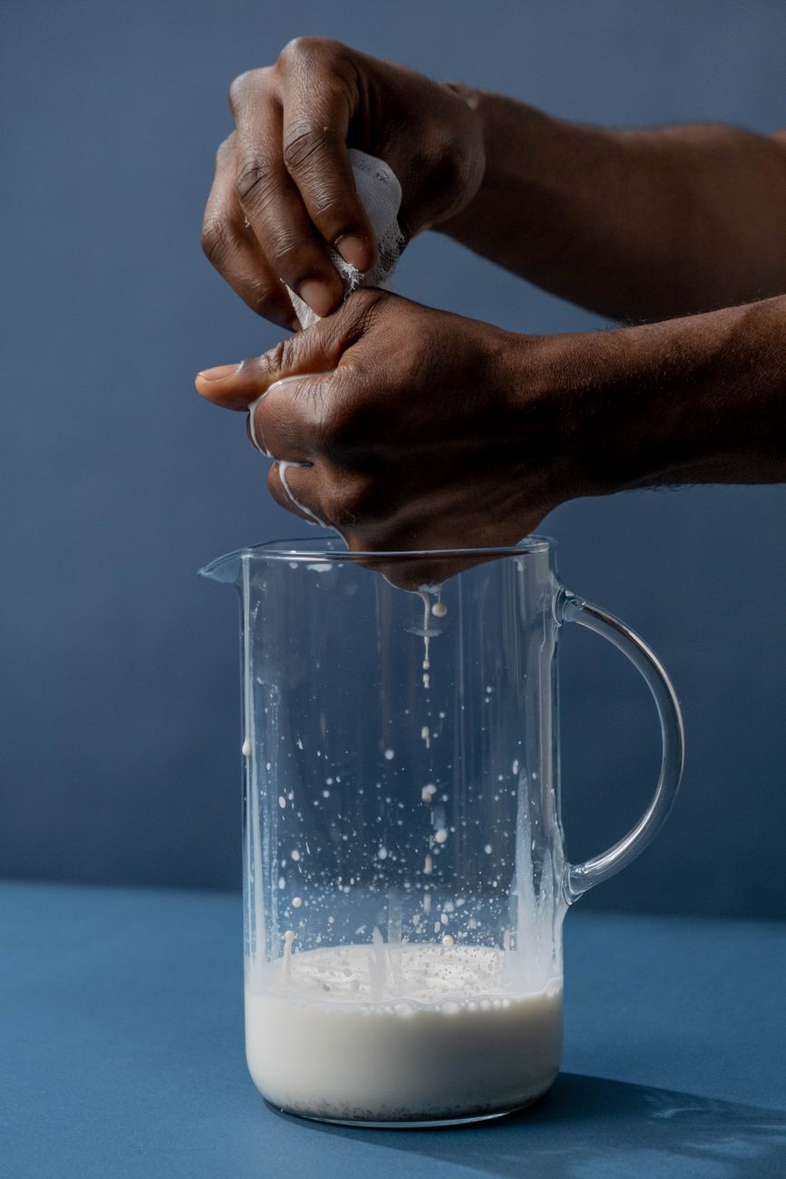 Gros plan de mains pressant un tissu pour filtrer du lait dans un bocal en verre sur fond bleu.