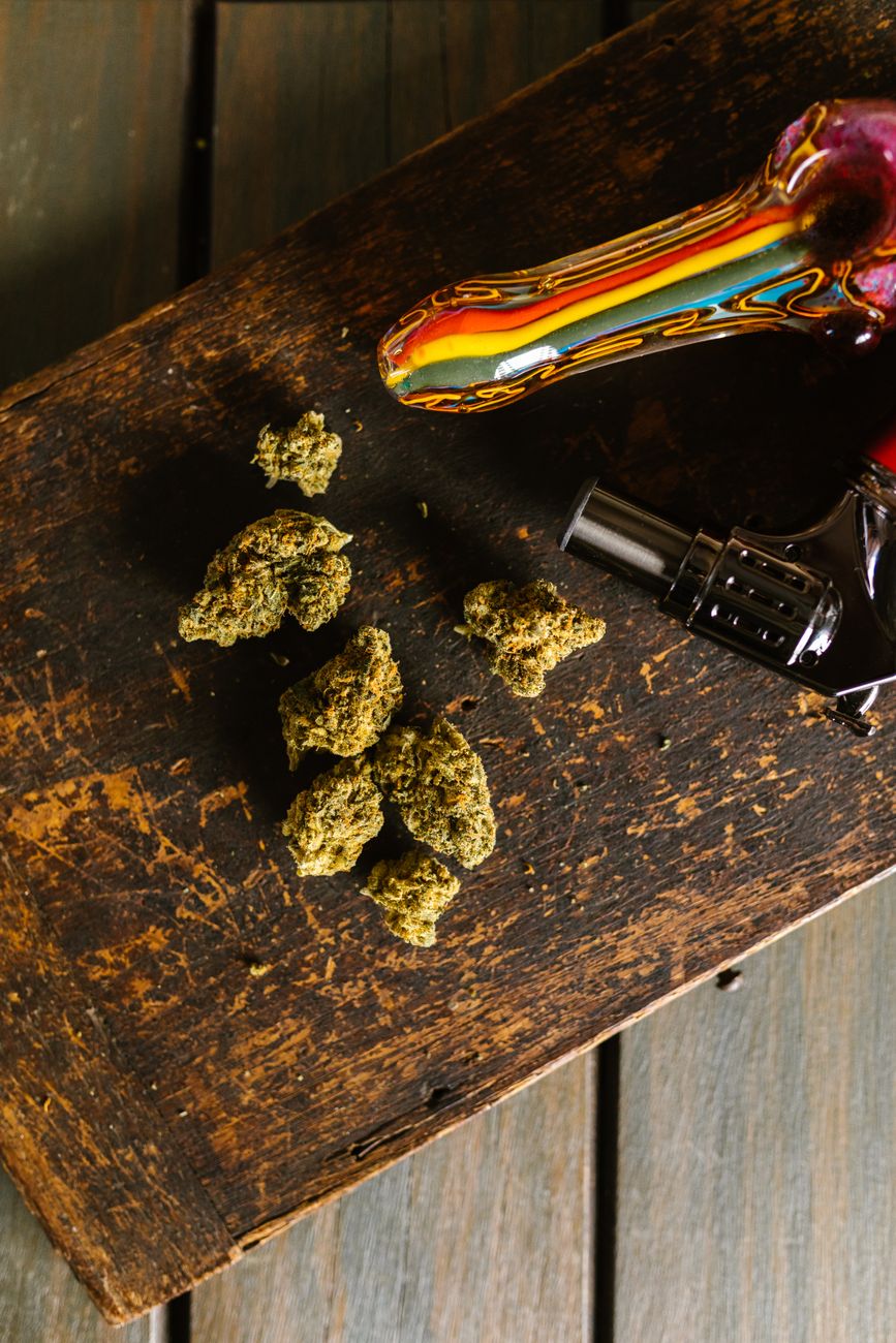 Une pipe en verre colorée et des bourgeons de cannabis posés sur une table en bois rustique en vue verticale.