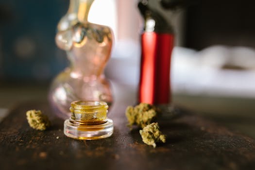 Bocal en verre contenant un concentré de cannabis, entouré de boutons de marijuana séchés et d'accessoires pour fumer sur une table.